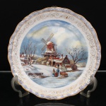 Prato decorativo executado em porcelana, esmaltação branca, aba perolada em tonalidade azul-lilás e acabamento iridescente, delimitada por filetes dourados. Medalhão central com decoração policroma representando paisagem rural de inverno, composta por moinho de vento, casario, árvores desfolhadas e figuras em caminho coberto de neve, sob céu azul com nuvens. Aba interna ornamentada por sequência de volutas estilizadas em dourado. Verso sem marca de manufatura. Medida: Diam.: 25,5 cm.