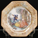 Porcelana Schmidt, Santa Catarina, Brasil (Fundada em 1945). Prato decorativo em porcelana, coleção Victorian Dreams  Pet and Toy Collection, Schmidt, segunda metade do século XX. Formato octogonal, aba decorada com efeito lustreware (iridescente/madrepérola), com padronagem marmorizada em tons de bronze, ocre e dourado, rematada por filete em douradura. Ao centro, reprodução policroma com cena galante de inspiração vitoriana, representando músico tocando instrumento de cordas diante de duas figuras femininas, em ambiente ajardinado, com fonte arquitetônica e composição floral. No verso, inscrições VICTORIAN DREAMS  Pet and toy COLLECTION e marca da manufatura SCHMIDT. Medida: Diam.: 26,5 cm.