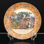 Prato de aparato executado em porcelana, decoração ao gosto Satsuma, Japão, período Showa. Ornamentação em esmaltes policromos sobre o vidrado, douração e aplicações em relevo pela técnica moriage. Ao centro, cena de gênero ambientada em jardim, com três figuras femininas em trajes tradicionais observando uma criança junto a um galo, ladeadas por pavão de cauda aberta, faisões, peônias e arvoredo; ao fundo, paisagem lacustre dominada pela silhueta do monte Fuji. A composição associa figuras, aves e florações ao repertório japonês de pássaros e flores(kach-ga). Aba em fundo ocre pontilhado, percorrida por composição alternada de flores, folhagens, cartelas e volutas azuis, com realces a ouro. Orla rematada por filete dourado. Reverso esmaltado em branco, sem marca de manufatura. Medida: Diam.: 30,5 cm.
