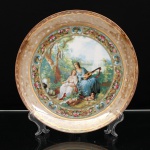 Prato decorativo executado em porcelana, aba decorada com efeito lustreware (iridescente/madrepérola), de padronagem marmorizada em tonalidades bege, champanhe e dourado, rematada por filetes dourados. Ao centro, reprodução policroma de composição de inspiração romântica, representando duas figuras femininas em ambiente campestre, uma delas tocando instrumento de cordas, acompanhadas por cão e cesta de flores, circundada por faixa ornamental composta por guirlanda de rosas e medalhões com urnas floridas, delimitada por filetes dourados. No verso, assinatura Bia. Medida: Diam.: 24,5cm.