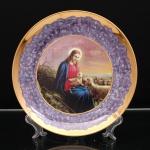 Prato decorativo executado em porcelana, aba decorada em fundo púrpura marmorizado, circundada por larga faixa dourada e rematada por filete na mesma tonalidade. Ao centro, reprodução policroma de composição religiosa representando Jesus Cristo como o Bom Pastor, sentado em paisagem campestre, com cordeiro ao colo e rebanho ao redor, iconografia alusiva à proteção e condução dos fiéis. Reserva central delimitada por filete dourado. No verso, assinatura manuscrita Bia. Medida: Diam.: 24,5cm.