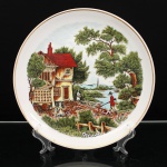 Porcelana Real, Mauá - SP(Fundada em 1943 por Fritz Erwin Schmidt). Prato decorativo executado em porcelana nacional, esmaltação branca, bordo contornado por friso em douradura. Ao centro, composição em estampagem policromada de caráter campestre, representando residência rural junto a curso dágua, figuras em atividades cotidianas e homem acompanhado por cães, tendo ao fundo vegetação e torre de igreja integradas à paisagem. Decoração em tonalidades verde, castanho, vermelho, azul e ocre. No verso, marca da manufatura em verde, constituída por coroa e inscrições "PORCELANA REAL - BRASIL". Medida: Diam.: 26,5cm.