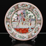 Prato de aparato executado em porcelana chinesa, estilo Cantão, decoração famille rose. Ornamentação em esmaltes policromos sobre o vidrado, com douração. Ao centro, cena de interior representando grupo de figuras femininas reunidas em torno de tabuleiro de Go(weiqi), entre cortinados, bambus, mobiliário e elementos arquitetônicos; uma das personagens porta leque, enquanto outra acompanha a partida. A cena integra o repertório das Quatro Artes da tradição letrada chinesa, no qual o weiqi figura ao lado da cítara qin, da caligrafia e da pintura. Aba compartimentada por reservas com aves, flores e frutos, alternadas com meandros e composições florais sobre fundo dourado; faixa externa em rosa com motivos estilizados. Orla rematada por filete a ouro. No verso, marca apócrifa Qianlong em vermelho-ferro, com a inscrição "Qianlong nian zhi", "Feito durante o reinado de Qianlong", alusiva ao reinado de Qianlong (1736-1795), dinastia Qing. Medida: Diam.: 27cm.