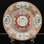Prato de aparato em porcelana chinesa, estilo Cantão, decoração famille rose sobre fundo millefleurs. Ornamentação em esmaltes policromos sobre o vidrado, com douração. Composição central em medalhão circular com flores, aves e borboletas, circundado por campo vermelho preenchido por florações azuis e volutas. Aba densamente ornamentada por ramagens floridas sobre fundo dourado, distribuída em reservas lobuladas com peônias, aves, borboletas e motivos vegetalistas, intercaladas por medalhões circulares com caracteres chineses de caráter auspicioso. A associação entre pássaros, flores e insetos deriva do repertório tradicional chinês de pássaros e flores(huaniao), recorrente nas artes decorativas. Orla rematada por filete a ouro. No verso, marca apócrifa Qianlong em vermelho-ferro, com a inscrição "Qianlong nian zhi", "Feito durante o reinado de Qianlong", alusiva ao reinado de Qianlong(1736-1795), dinastia Qing. Medida: Diam.: 27cm.