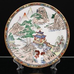 Prato de aparato em porcelana chinesa, decoração famille rose. Ornamentação em esmaltes policromos sobre o vidrado, com douração. Ao centro, paisagem montanhosa de inspiração shan shui, composta por formações rochosas, pinheiros, salgueiro, cursos dágua e pavilhão sobre as águas, ligado à margem por passadiço, com figuras em seu interior. Ao longe, picos montanhosos emergem entre planos sucessivos da paisagem. Aba estreita ornamentada por sequência de flores, frutos e folhagens sobre fundo dourado, delimitada internamente por friso geométrico. Bordo contornado por filete a ouro. No verso, marca apócrifa Qianlong em vermelho-ferro, com a inscrição "Qianlong nian zhi", "Feito durante o reinado de Qianlong", alusiva ao reinado de Qianlong(1736-1795), dinastia Qing.Medida: Diam.: 27cm.