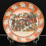 Prato de aparato em porcelana japonesa, decoração ao gosto Satsuma, Japão, período Showa. Ornamentação em esmaltes policromos sobre o vidrado, douração e aplicações em relevo pela técnica moriage. Ao centro, cena de gênero representando grupo de figuras femininas e crianças em passeio por jardim, trajando quimonos de padrões diversos, entre pinheiros, bambuzais, florações e formações rochosas. Ao fundo, paisagem lacustre articulada por ponte arqueada, pavilhões e elevações montanhosas. A composição remete ao repertório das bijin-ga, representações femininas difundidas na pintura e na gravura japonesa. Aba em fundo de tonalidade coral pontilhado, ornamentada por reservas com motivos florais e folhagens, entremeadas por arabescos dourados. Orla rematada a ouro. Reverso esmaltado em branco, sem marca de manufatura aparente. Medida: Diam.: 30,5cm.