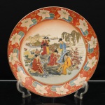 Prato de aparato executado em porcelana japonesa, decoração ao gosto Satsuma, Japão, período Showa. Ornamentação em esmaltes policromos sobre o vidrado, douração e aplicações em relevo pela técnica moriage. Ao centro, cena de gênero de inspiração bijin-ga, representando grupo de mulheres em trajes tradicionais reunidas às margens de um regato, entre crisântemos, peônias e cerejeira em flor; duas figuras percorrem a paisagem ao fundo, onde se sucedem bambuzal, salgueiro, casario, embarcações, ponte arqueada, pavilhão e formações montanhosas. A composição evoca o repertório japonês de passeios e encontros femininos em jardins, associado à contemplação sazonal da natureza. Aba em fundo de tonalidade coral, ornamentada por reservas florais sobre campos azuis, entre folhagens e arabescos dourados. Orla rematada por filete a ouro. Reverso esmaltado em branco, sem marca de manufatura aparente. Medida: Diam.: 30,5cm.