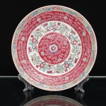 Prato decorativo em porcelana chinesa, famille rose, no estilo Mun Shou, China, século XX. Decoração policromada sobre esmalte, organizada em campos concêntricos, com reservas florais sobre fundo rosa, preenchido por motivos estilizados, e friso externo de meandros em azul, rosa e amarelo. A ornamentação reúne símbolos auspiciosos da tradição chinesa, entre eles flores de ameixeira, crisântemos, pêssegos, morcegos e motivos ruyi, associados à longevidade, felicidade, prosperidade e realização dos bons desejos; o friso de meandros remete à continuidade e perpetuidade. No reverso, marca ST, acompanhada dos caracteres Zhongguo zhizao, Fabricado na China, e da inscrição Made in China. Apresenta suporte metálico para suspensão adaptado. Medida: Diam.: 23cm.