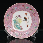 Prato de aparato em porcelana chinesa, decoração famille rose. Jingdezhen. Ornamentação em esmaltes policromos sobre o vidrado. Ao centro, reserva central representando ave pousada entre ramagens floridas, crisântemos e peônias, delimitada por contorno recortado em vermelho-ferro. Aba em fundo rosa, preenchida por motivos ruyi e flores estilizadas, entremeados por ramos de peônias e folhagens. A composição reúne símbolos auspiciosos da tradição chinesa, associados à longevidade, prosperidade, renovação, boa fortuna e realização dos desejos. Borda delimitada por filete em vermelho-ferro. No verso, marca "Zhongguo Jingdezhen", "Jingdezhen, China". Medida: Diam.: 20,5cm.