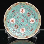 Prato decorativo em porcelana chinesa, estilo famille rose, Jingdezhen. Ornamentação policromada sobre fundo turquesa, estruturada por quatro reservas circulares com caracteres chineses em vermelho-ferro, entre motivos florais, ramagens e emblemas auspiciosos. Destaca-se o caractere(shou), símbolo de longevidade, integrado a um repertório ornamental associado a votos de longevidade, prosperidade e boa fortuna. Campo preenchido por motivos estilizados de nuvens, tradicionalmente relacionados à auspiciosidade e às bênçãos celestes, intercalados por flores e elementos fitomórficos. Aba delimitada por faixa de grega chinesa(leiwen) policromada. No reverso, marcado com a inscrição "Zhongguo Jingdezhen", "Made in China", indicando produção de Jingdezhen, tradicional centro de manufatura de porcelana na província de Jiangxi, China. Apresenta suporte metálico adaptado para suspensão no verso. Medida: Diam.: 20,5 cm.