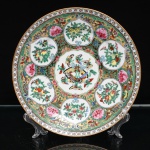 Prato decorativo em porcelana chinesa, estilo famille rose. Ornamentação policromada e dourada, organizada em reservas lobuladas sobre campo densamente preenchido por ramagens, flores, frutos e borboletas. Ao centro, composição com borboletas de diferentes espécies, motivo tradicional associado à felicidade, longevidade e união conjugal. Reservas periféricas apresentam frutos e flores, entre eles pêssegos, romãs e frutos cítricos, vinculados na simbologia chinesa à longevidade, fertilidade, descendência e prosperidade. Grandes flores rosadas, ramagens e folhagens complementam a ornamentação sobre fundo dourado. Aba delimitada por faixa geométrica reticulada com pequenas flores estilizadas, rematada por filete dourado. No verso, marca sigilar em vermelho-ferro com caracteres chineses, parcialmente encoberta por etiqueta, além de suporte metálico para suspensão(adaptado). Medida: Diam.: 23 cm.