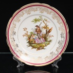 Porcelana Real, Mauá - SP(Fundada em 1943 por Fritz Erwin Schmidt). Prato decorativo em porcelana nacional, esmaltagem vítrea na cor branca, aba recortada e bordo realçado por filete dourado. Orla ornamentada por faixa em tonalidade vinho, delimitada por frisos dourados, e composição contínua de arabescos, elementos florais e reservas reticuladas em ouro. Ao centro, cena galante em estampagem policroma com paisagem campestre, representando figura feminina reclinada junto a cesto, acompanhada por figura masculina tocando instrumento de sopro, entre vegetação e arvoredo. No reverso, marca da manufatura "Real - S. Paulo - Made in Brazil. Medida: Diam.: 24,5cm.