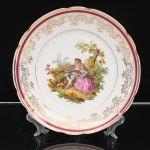 Porcelana Real, Mauá - SP(Fundada em 1943 por Fritz Erwin Schmidt). Prato decorativo em porcelana nacional, esmaltagem vítrea na cor branca, aba recortada e bordo realçado por filete dourado. Orla ornamentada por faixa em tonalidade vinho, delimitada por frisos dourados, e composição contínua de arabescos, elementos florais e reservas reticuladas em ouro. Ao centro, cena galante em estampagem policromada de caráter pastoril, representando casal em ambiente campestre, com figura masculina apresentando pequena gaiola à figura feminina sentada, acompanhados por cão, entre arvoredo e vegetação. No verso, marca da manufatura "Real - S. Paulo - Made in Brazil. Medida: Diam.: 24,5 cm.