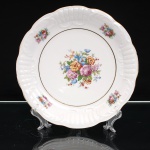 Porcelana Schmidt, Santa Catarina, Brasil(Fundada em 1945). Prato decorativo em porcelana nacional, esmaltagem vítrea na cor branca, aba moldada em relevo com reservas alternadas de concheados e caneluras, bordo ondulado rematado por filete dourado. Campo central delimitado por friso em ouro e ornamentado com composição floral policroma, reunindo rosas e flores estilizadas em tonalidades rosa, azul, amarelo, alaranjado e verde; quatro ramalhetes menores distribuídos pela aba. No verso, marca em verde "PORCELANA SCHMIDT  POMERODE - S. CATARINA". Medida: Diam.: 25,5cm.