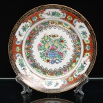 Prato decorativo em porcelana chinesa, estilo Cantão, paleta famille rose. Decoração policromada e dourada. Ao centro, composição de flores dispostas em vaso, associadas a frutos e borboletas. Na tradição ornamental chinesa, as borboletas relacionam-se à felicidade, longevidade e união conjugal; os frutos, à abundância e prosperidade; e as flores, conforme a espécie representada, integram o repertório auspicioso ligado à renovação, longevidade e prosperidade. A composição é circundada por faixa de reservas com motivos florais, frutos, aves e borboletas, intercaladas sobre fundo dourado com ramagens. Aba externa organizada em reservas sobre fundo vermelho, contendo flores, frutos e pássaros, delimitada por larga faixa negra com arabescos dourados e filete dourado no bordo. No reverso, marca impressa em vermelho-ferro com as inscrições DECORATIVE HAND PAINTING / MADE IN CHINA, acompanhadas de caracteres chineses. Peça provida posteriormente de suporte metálico para suspensão(adaptado). Medidas: Diam.: