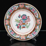 Prato decorativo em porcelana chinesa, paleta famille rose, China, século XX. Prato circular em porcelana branca, com decoração policromada e dourada. Ao centro, vaso com arranjo de peônias e flores de ameixeira, ladeado por pêssegos e bambu. Na tradição chinesa, a peônia associa-se à prosperidade e distinção; a flor de ameixeira, à perseverança e renovação; o pêssego, à longevidade; e o bambu, à retidão e resistência. Aba ornamentada sobre fundo dourado, dividida em reservas contendo flores, pêssegos e motivos vegetais, alternadas com medalhões do caractere(fú), símbolo de felicidade, boa fortuna e bênçãos. Filetes dourados delimitam a composição e o bordo. No reverso, marca ST acompanhada dos caracteres "Zhongguo zhizao", "fabricado na China", e da inscrição MADE IN CHINA. Peça provida posteriormente de suporte metálico para suspensão(adaptado). Medida: Diam.: