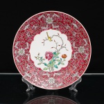 Prato decorativo em porcelana chinesa, paleta famille rose, Jingdezhen. Ornamentação em esmaltes policromos. Reserva central recortada contendo composição de roseiras floridas, com ave pousada sobre ramo. Na simbologia chinesa, a rosa associa-se à juventude, beleza e continuidade das estações, enquanto a representação de flores e pássaro integra o tradicional gênero huaniao, flores e aves, relacionado à harmonia do mundo natural. Aba revestida em esmalte rosa-carmim, preenchida por motivos curvilíneos repetidos e quatro composições florais dispostas simetricamente, com detalhes em verde, branco e vermelho-ferro. Bordo delimitado por filete em vermelho-ferro. No reverso, marca em vermelho-ferro "Zhongguo Jingdezhen", "Jingdezhen, China", acompanhada da inscrição "MADE IN CHINA" e do número 06. Jingdezhen, na província de Jiangxi, constitui um dos principais centros históricos da produção de porcelana chinesa. Peça provida posteriormente de suporte metálico para suspensão(adaptado). Medida: Diam.: