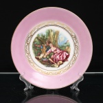 Porcellana Mauá, Mauá - SP(Fundada em 1937 por Fritz Erwin Schmidt / ativa até 1968). Prato de coleção em porcelana nacional, aba circundada por faixa de tonalidade rosa, delimitada por filete dourado no bordo. Centro ornado por faixa de arabescos estilizados em dourado sobre fundo branco, circundando reserva central policroma com cena galante de inspiração rococó, representando casal em ambiente campestre, acompanhado de caprino e elementos florais. Na composição figura a inscrição Boucher - Musée du Louvre - Amsterdam-Holland, indicando reprodução de motivo atribuído a François Boucher(1703-1770), pintor francês associado ao Rococó. No verso, marca estampada "PORCELLANA MAUÁ" e suporte metálico para suspensão. Medida: Diam.: