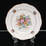Porcelana Schmidt, Santa Catarina, Brasil (Fundada em 1945). Prato decorativo em porcelana nacional, esmaltagem vítrea na cor branca, aba moldada em relevo com segmentos canelados e bordo ondulado, rematado por filete dourado. Ao centro, composição floral policroma formada por rosas, flores e folhagens em tonalidades de rosa, azul, amarelo, alaranjado e verde; motivos florais menores distribuídos pela aba. Cavado delimitado por filete dourado. No verso, marca da manufatura estampada em verde, com as inscrições "PORCELANA SCHMIDT / P. INDUSTRIA / CATARINA". Apresenta dispositivo metálico para suspensão. Medida: Diam.: