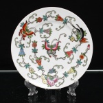 Prato de coleção em porcelana chinesa, da região de Jingdezhen, decorado em esmaltes policromos, com composição formada por borboletas entre ramagens sinuosas, flores e frutos, distribuída de maneira assimétrica pela superfície. Ao centro, borboleta de asas abertas em tons de rosa, verde e azul; ao redor, exemplares menores associados a flores campanuladas e frutos. O repertório ornamental reúne motivos tradicionais chineses de caráter auspicioso. A borboleta(húdié) relaciona-se à longevidade, felicidade e união conjugal; sua associação com flores reforça a ideia de harmonia e prosperidade. Frutos e flores integram o vocabulário simbólico ligado à fertilidade, abundância e continuidade das gerações. As ramagens contínuas que percorrem a composição remetem ao conceito de perpetuidade e sucessão. No verso, marca em vermelho-ferro "Zhongguó Jingdézhèn", "Jingdezhen, China", acompanhada da inscrição "MADE IN CHINA" e da letra N. Jingdezhen, na província de Jiangxi, constitui um dos principais centros históricos de produção de porcelana da China. Peça provida posteriormente de suporte metálico para suspensão(adaptado). Medida: Diam.: