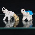 Três(03) figuras de elefantes em cerâmica, século XX. Três representações modeladas com a tromba elevada, em diferentes dimensões. Exemplares com acabamentos distintos: um revestido por esmalte branco, com apontamentos dourados e castanhos; outro em tonalidade marfim, com detalhes em azul e dourado; o terceiro em branco, com manta azul-turquesa sobre o dorso, traços dourados, amarelos, vermelhos e pretos. Medidas: