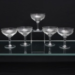 Conjunto de cinco(05) taças em meio cristal incolor,bojo em formato de coupe,  dotado de ornamentação executada em gravação, organizada em faixas concêntricas: friso superior de elementos lanceolados sucessivos e, abaixo, composição contínua de volutas, círculos e entrelaçamentos geométricos. Medidas: Alt.: x Diam.: