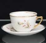 Xícara para chá em porcelana, esmaltagem vítre na cor branca, bojo hemisférico, com alça lateral estilizada e filetagem dourada no bordo. Superfície ornada por composição botânica em estampagem policromada, constituída por flores e ramagens em tons de branco, bege, verde e vermelho. Pires circular com cavidade central delimitada por filete dourado, repetindo o repertório floral da xícara e arremate em ouro na borda. Alça integralmente dourada. Medidas: Xícara: Alt.: 5,5 x Diam.: 8,5 ; Pires: Diam.: 13,5 cm.