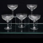 Conjunto de cinco(05) taças em meio cristal incolor,bojo em formato de coupe,  dotado de ornamentação executada em gravação, organizada em faixas concêntricas: friso superior de elementos lanceolados sucessivos e, abaixo, composição contínua de volutas, círculos e entrelaçamentos geométricos. Medidas: Alt.: x Diam.: