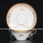 Porcelana Real, Mauá - SP(Fundada em 1943 por Fritz Erwin Schmidt). Xícara para chá em porcelana nacional, esmaltagem vítrea na cor branca, bojo obcônico assente sobre base circular, alça lateral em voluta, acompanhada de pires circular com cavidade central para encaixe. Decoração em douradura, constituída por friso contínuo junto ao bordo, faixa ornamental de inspiração neoclássica formada por elementos fitomórficos repetidos e filetação no pires. Apresenta as inscrições Você, na face da xícara, e Eu, na aba do pires, ambas em caligrafia dourada. Marca da manufatura no verso. Medidas: Xícara: Alt.: 4,5 x Comp.: 11,5 x Larg.: 10cm. ; Pires: Diam.: 14cm.