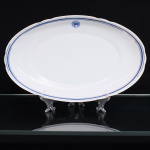 Porcelana Real, Mauá - SP(Fundada em 1943 por Fritz Erwin Schmidt). Travessa de serviço do Lloyd Brasileiro em porcelana nacional, formato oval, esmaltação branca, decoração composta por faixa azul cobalto entre filetes dourados, acompanhando o perímetro. No verso, emblema do Lloyd Brasileiro, com bandeira da companhia ao centro, e marca da manufatura Real  S. Paulo  Made in Brazil. Peça integrante de serviço de mesa institucional produzido pela Porcelana Real para o Lloyd Brasileiro, extinta empresa de navegação incorporada ao patrimônio nacional e constituída como autarquia federal em 1937. Medidas: Comp.: x Larg.: