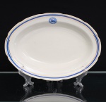 Porcelana D.Pedro II, Rio de Janeiro - RJ(Fundada em 1921 por Tinoco Machado, ativa até a década de 1980). Travessa de serviço do Lloyd Brasileiro em porcelana nacional, formato oval, esmaltação branca, decoração composta por faixa azul cobalto entre filetes dourados, acompanhando o perímetro. No verso, emblema do Lloyd Brasileiro, com bandeira da companhia ao centro, e marca da manufatura. Peça integrante de serviço de mesa institucional produzido pela Porcelana Real para o Lloyd Brasileiro, extinta empresa de navegação incorporada ao patrimônio nacional e constituída como autarquia federal em 1937. Medidas: Comp.: x Larg.: