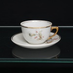 PORCELANA RAMI, Jundiaí São Paulo (fundada em 1930). Xícara para chá em porcelana, esmaltagem vítre na cor branca, bojo hemisférico, com alça lateral estilizada e filetagem dourada no bordo. Superfície ornada por composição botânica em estampagem policromada, constituída por flores e ramagens em tons de branco, bege, verde e vermelho. Pires circular com cavidade central delimitada por filete dourado, repetindo o repertório floral da xícara e arremate em ouro na borda. Alça integralmente dourada. Medidas: Xícara: Alt.: 5,5 x Diam.: 8,5 ; Pires: Diam.: 13,5 cm.