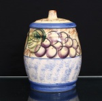 Pote com tampa em cerâmica esmaltada, decoração frutífera policroma. Corpo de perfil abaulado, assente sobre base circular, com faixa inferior em azul e campo central de fundo branco azulado. Faixa superior ornamentada com composição de frutos e folhagens, incluindo cachos de uvas e frutos de formas variadas, executados em verde, amarelo, laranja, violeta e castanho, definidos por contornos em tonalidade escura. Tampa ligeiramente abaulada, dotada de puxador cilíndrico, com acabamento esponjado em castanho sobre fundo creme e faixa azul junto ao encaixe. Decoração executada à mão. Sem marca de manufatura. Medidas: Alt.: 24,5 x Diam.: 18cm.