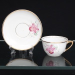 PORCELANA RAMI, Jundiaí  São Paulo (fundada em 1930). Xícara para chá em porcelana nacional, de esmaltação branca, decoração floral em estampagem policromada, representando orquídeas em tonalidades rosa, verde e amarelo. Bojo hemisférico assente sobre base circular, com alça lateral angular realçada por douradura; pires circular de aba ligeiramente côncava, ornamentado pelo mesmo motivo botânico. Bordas e cavidade de assentamento delimitadas por frisos dourados. Marca de manufatura PORCELANA RAMI na base. Medidas: Xícara: Alt.: 5 x Comp.: 10,5 x Larg.: 8,5cm. ; Pires: Diam.: 14cm.