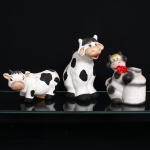 Três(03) peças utilitárias e decorativas em cerâmica, com representação zoomórfica de bovinos. Composto por duas figuras modeladas em forma de vaca, uma delas configurada como recipiente, e peça de maior porte com cavidade superior e corpo integrado à representação do animal. Superfícies esmaltadas em branco, com manchas pretas e detalhes policromos em bege, rosa, vermelho, amarelo e azul. Cabeças modeladas com focinho proeminente, orelhas, chifres e feições delineadas à mão. Uma das peças conserva aplicação de laço em tecido vermelho junto ao pescoço. Decoração de caráter lúdico, associada à produção de cerâmica utilitária doméstica. Sem marca de manufatura. Medidas: Alt.: 26 x Comp.: 27 x Larg.: 14,5 cm; Alt.: 19,5 x Comp.: 18 x Larg.: 13,5 cm; Alt.: 13,5 x Comp.: 23,5 x Larg.: 10 cm.