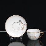 PORCELANA RAMI, Jundiaí São Paulo (fundada em 1930). Xícara para chá em porcelana, esmaltagem vítre na cor branca, bojo hemisférico, com alça lateral estilizada e filetagem dourada no bordo. Superfície ornada por composição botânica em estampagem policromada, constituída por flores e ramagens em tons de branco, bege, verde e vermelho. Pires circular com cavidade central delimitada por filete dourado, repetindo o repertório floral da xícara e arremate em ouro na borda. Alça integralmente dourada. Medidas: Xícara: Alt.: 5,5 x Diam.: 8,5 ; Pires: Diam.: 13,5 cm.