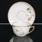 PORCELANA RAMI, Jundiaí São Paulo (fundada em 1930). Xícara para chá em porcelana, esmaltagem vítre na cor branca, bojo hemisférico, com alça lateral estilizada e filetagem dourada no bordo. Superfície ornada por composição botânica em estampagem policromada, constituída por flores e ramagens em tons de branco, bege, verde e vermelho. Pires circular com cavidade central delimitada por filete dourado, repetindo o repertório floral da xícara e arremate em ouro na borda. Alça integralmente dourada. Medidas: Xícara: Alt.: 5,5 x Diam.: 8,5 ; Pires: Diam.: 13,5 cm.