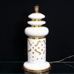 Cerâmica Artística Tupy Ltda., São Caetano do Sul, Brasil. Abajur de mesa executado em cerâmica esmaltada e metal dourado, c.1980. Corpo em cerâmica de esmaltação branca, estruturado em base circular, fuste cilíndrico vazado por motivos geométricos e curvilíneos, bojo anelar e coroamento escalonado. Elementos metálicos em acabamento dourado delimitam a base e as diferentes seções do corpo. Montagem elétrica interna, com soquete superior para cúpula e lâmpada. Conserva etiqueta original da Cerâmica Artística Tupy Ltda., com endereço à Rua Oswaldo Cruz, 162, São Caetano do Sul, São Paulo. Medidas: Alt.: x Diam.: