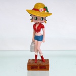 BRINQUEDO / COLECIONISMO. Estatueta de coleção executada em resina policromada, figurando a personagem Betty Boop em trajes de camponesa / fazendeira. Coleção oficial licenciada, final do século XX ou início do século XXI. Personagem representada em pé sobre base retangular que simula solo de terra batida, apresenta-se com sua fisionomia característica de traços marcantes, grandes olhos verdes e brincos de argola amarelos. Traja camisa vermelha com amarração frontal revelando o abdômen, shorts jeans curto azul(hot pants), meias brancas e botas de cano curto na cor vermelha. Sobre a cabeça, ostenta grande chapéu de palha texturizado, ornado por fita vermelha com laço posterior e arranjo lateral composto por folhas verdes e flores em tons de rosa, vermelho e branco. A figura segura com a mão direita um galão de leite metálico prateado com alça. Na face frontal da base, inscrição em relevo com o nome da personagem "BETTY BOOP", destacado em esmalte vermelho com contorno preto. No reverso da base, apresenta inscrição de direitos autorais e licenciamento oficial gravada: "KFS/FS TM Hearst/FS"(King Features Syndicate e Fleischer Studios, distribuído pela Hearst Holdings). Medidas: Alt.: x Comp.: x Larg.: