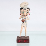 BRINQUEDO / COLECIONISMO. Estatueta de coleção executada em resina policromada, figurando a personagem Betty Boop vestida como Chef de Cozinha. Coleção oficial licenciada, final do século XX ou início do século XXI. Apresenta-se em posição frontal, com seus característicos olhos verdes e brincos de argola na cor vermelha. Traja uniforme clássico de mestre-cuca(dólmã) branco, debruns e botões em azul, modelagem curta, complementado por um avental amarrado por laço azul em seu reverso. Sobre a cabeça, ostenta um alto chapéu de chef(toque blanche) texturizado, decorado na base frontal pela inscrição manuscrita "Chef Betty"; segura com a mão esquerda uma panela de cabo longo em tom marrom-terroso, enquanto leva à boca uma colher prateada com a mão direita, simulando degustação. Calça sapatos de salto alto clássicos na cor branca. Na face frontal da base, inscrição em relevo com o nome da personagem "BETTY BOOP", destacado em esmalte vermelho com contorno preto. No reverso da base, apresenta inscrição de direitos autorais e licenciamento oficial gravada: "KFS/FS TM Hearst/FS"(King Features Syndicate e Fleischer Studios, distribuído pela Hearst Holdings). Medidas: Alt.: x Comp.: x Larg.: