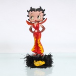 BRINQUEDO / COLECIONISMO. Estatueta de coleção executada em resina policromada, figurando a personagem Betty Boop em trajes estilo diabete / vamp. Distribuição oficial licenciada pela Editora Salvat do Brasil Ltda.(Edição n.20). A icônica personagem surge representada em pé, em pose altiva com as mãos na cintura, ostentando seus tradicionais olhos verdes e brincos de argola dourados. Traja macacão justo na cor vermelha com abertura circular na região do abdômen e calça estilo bailarina(boca de sino), ricamente ornamentado por estampas de labaredas/chamas amarelas e laranjas nas pernas. O traje é complementado por uma imponente gola alta pontiaguda e luvas longas que cobrem até os cotovelos, recobertas por acabamento em purpurina / glitter vermelho de forte efeito visual. Calça sapatos de salto alto na cor vermelha, assentada sobre base circular preta, guarnecida e decorada em todo o seu contorno por plumas ou penugem sintética na cor preta. Na face frontal da base, destaca-se medalhão amarelo em formato de labareda com a inscrição impressa em inglês: "It's so Good to be so Bad!" ("É tão bom ser tão má!"). No verso da base, apresenta inscrição de direitos autorais desgastada e, sob o fundo da base, etiqueta comercial com dados completos de importação, CNPJ da distribuidora e a especificação técnica: "Atenção! Artigo para colecionistas. Não é um brinquedo". Medidas: Alt.: x Comp.: x Larg.: 