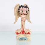 BRINQUEDO / COLECIONISMO. Estatueta decorativa colecionável em resina policromada figurando a personagem Betty Boop em trajes de Anjo. Coleção oficial licenciada fabricada pela Vandor(Salt Lake City, UT), datada de 2001. A icônica personagem surge representada em posição frontal com as mãos unidas delicadamente à frente do corpo, ostentando seus tradicionais olhos verdes e brincos de argola dourados. Traja longo vestido de gala em tom marfim/creme, acinturado por fino cordão vermelho com amarração em laço no reverso. A saia, de caimento fluido estilo sereia, abre-se em ampla nesga em sua base revelando forro interior contrastante na cor vermelha, sapatilhas de amarração trançada e pés assentados sobre uma nuvem estilizada. Em suas costas, projeta-se um par de grandes asas de anjo texturizadas e totalmente recobertas por acabamento em purpurina / glitter dourado de forte efeito decorativo. A figura encontra-se fixada sobre base circular marfim, guarnecida e decorada em todo o seu contorno por plumas ou penugem na cor bege-claro / champanhe. Na face frontal da base, destaca-se medalhão azul-celeste em formato de nuvem com a inscrição impressa em inglês: "It's not easy to be this Good!" ("Não é fácil ser tão boa!"). Sob o fundo da base, apresenta carimbo oficial de licenciamento da marca "BETTY BOOP" datado de 2001, com créditos à King Features Syndicate, Inc. / Fleischer Studios, Inc. / Hearst Holdings, Inc., além da indicação "Handmade in China - Vandor - SLC, UT", parcialmente coberta por resquício de etiqueta adesiva. Medidas: Alt.: x Comp.: x Larg.: