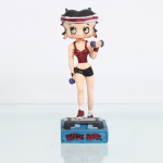 BRINQUEDO / COLECIONISMO. Estatueta decorativa colecionável em resina policromada figurando a personagem Betty Boop Praticando Fitness / Aeróbica. Distribuição oficial licenciada pela Editora Salvat do Brasil Ltda. (Edição nº 22). A icônica personagem surge representada em ação, executando exercícios físicos sobre uma plataforma de step cinza e preta. Apresenta-se com seus característicos grandes olhos verdes e brincos de argola na cor vermelha, trazendo a cabeça cingida por uma faixa de suor(headband) nas cores vermelha e branca. Traja conjunto esportivo composto por top de alças em tom vermelho-grená metálico e shorts curto(hot pants) na cor preta. Segura um par de halteres(pesos) azuis, um em cada mão, elevando o braço esquerdo em flexão. Calça meias brancas de cano médio e tênis esportivos vermelhos e brancos.  Fixada sobre uma base retangular texturizada azul-turquesa. Na face frontal da base, inscrição em relevo com o nome da personagem "BETTY BOOP", destacado em esmalte vermelho com contorno preto. No reverso da base, apresenta inscrição de direitos autorais e licenciamento oficial gravada: "KFS/FS TM Hearst/FS". Sob o fundo da base, apresenta etiqueta comercial completa de importação com dados da distribuidora nacional, CNPJ e a indicação técnica da coleção: "For edition 22 - Artigo para colecionistas. Não é um brinquedo". Medidas: Alt.: x Comp.: x Larg.: