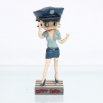 BRINQUEDO / COLECIONISMO. Estatueta decorativa colecionável em resina policromada figurando a personagem Betty Boop vestida como Policia. Distribuição oficial licenciada pela Editora Salvat do Brasil Ltda. A icônica personagem surge representada em pé sobre base retangular cinza que simula uma faixa de pedestres zebrada. Apresenta-se em posição frontal, prestando continência com a mão direita elevada à têmpora, ostentando seus tradicionais olhos verdes e brincos de argola na cor amarela. Traja uniforme clássico de guarda policial composto por camisa de mangas curtas em tom azul-claro com gola aberta, saia justa estilo minissaia em azul-marinho, cinto com fivela prateada e algemas metálicas penduradas na lateral esquerda da cintura. Sobre a cabeça, ostenta quepe policial azul com aba preta e insígnia de estrela/escudo na cor azul-claro. Calça sapatos de salto alto na cor azul-marinho.Na face frontal da base, inscrição em relevo com o nome da personagem "BETTY BOOP", destacado em esmalte vermelho com contorno preto. No reverso da base, apresenta inscrição de direitos autorais e licenciamento oficial gravada: "KFS/FS TM Hearst/FS". Medidas: Alt.: x Comp.: x Larg.: