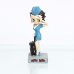 BRINQUEDO / COLECIONISMO. Estatueta decorativa colecionável em resina policromada figurando a personagem Betty Boop vestida como Comissária de Bordo / Aeromoça. Coleção oficial licenciada. A icônica personagem surge representada em pé sobre base retangular cinza, simulando o piso de um terminal aeroportuário. Apresenta-se em pose clássica, prestando uma saudação com a mão esquerda enluvada, enquanto ostenta seus característicos olhos verdes e brincos de argola na cor amarela. Traja uniforme retrô de tripulação de cabine composto por tailleur acinturado de mangas curtas e saia justa em tom azul-celeste, realçado por abotoamento frontal dourado e gola de camisa branca sobreposta. Sobre a cabeça, ostenta casquete(garrison cap) no mesmo tom azul do uniforme. Com a mão direita, segura a alça estendida de uma mala de viagem de rodinhas na cor preta, posicionada lateralmente. Calça sapatos de salto alto clássicos também em azul-celeste.Na face frontal da base, inscrição em relevo com o nome da personagem "BETTY BOOP", destacado em esmalte vermelho com contorno preto. No reverso da base, apresenta inscrição de direitos autorais e licenciamento oficial gravada: "KFS/FS TM Hearst/FS". Medidas: Alt.: x Comp.: x Larg.: