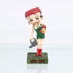 BRINQUEDO / COLECIONISMO. Estatueta decorativa colecionável em resina policromada figurando a personagem Betty Boop vestida como Jardineira / Camponesa. Coleção oficial licenciada, final do século XX ou início do século XXI. A icônica personagem surge representada em pé sobre base retangular texturizada em tom verde-musgo simulando gramado. Apresenta-se com o corpo levemente inclinado à frente, ostentando seus tradicionais grandes olhos verdes e brincos de argola amarelos. Traja jardineira jeans curta na cor verde, sobreposta a uma blusa regata branca, complementada por lenço amarrado na cabeça(bandana) na cor vermelha com nó em seu reverso. Com a mão direita voltada para baixo, segura um regador de plantas metálico estilizado na cor cinza-escura; sob o braço esquerdo, carrega uma cesta de vime preenchida por hortaliças e flores policromadas em tons de vermelho, verde e laranja. Calça galochas de cano médio na cor vermelha. Na face frontal da base, inscrição em relevo com o nome da personagem "BETTY BOOP", destacado em esmalte vermelho com contorno preto. No reverso da base, apresenta inscrição de direitos autorais e licenciamento oficial gravada: "KFS/FS TM Hearst/FS". Medidas: Alt.: x Comp.: x Larg.: cm.
