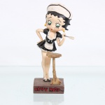 BRINQUEDO / COLECIONISMO. Estatueta decorativa / boneca colecionável em resina policromada figurando a personagem Betty Boop vestida como "Camareira Francesa"(French Maid). Coleção oficial licenciada, final do século XX ou início do século XXI. A icônica personagem surge representada em pose inclinada para o lado esquerdo, ostentando seus tradicionais olhos verdes e brincos de argola amarelos. Traja clássico uniforme de camareira composto por vestido curto preto com decote profundo, mangas bufantes adornadas com babados brancos e um pequeno avental de babados brancos amarrado na cintura por um laço em seu reverso. Sobre a cabeça, traz uma tiara ou casquete de babados combinando em tons de branco e preto. Com a mão esquerda elevada, segura um espanador de pó estilizado na cor marfim; o braço direito apoia-se na cintura, enquanto o corpo se projeta parcialmente atrás de uma banqueta alta ou mesa de apoio redonda de madeira. Calça sapatos clássicos de salto alto no modelo peep toe em tons de preto e branco. Figura encontra-se fixada sobre base retangular de tonalidade marrom. Na face frontal da base, inscrição em relevo com o nome da personagem "'BETTY BOOP", destacado em esmalte vermelho com contorno preto. No reverso da base, apresenta inscrição de direitos autorais e licenciamento oficial gravada: "KFS/FS TM Hearst/FS". Medidas: Alt.: x Comp.: x Larg.: