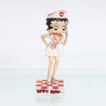 BRINQUEDO / COLECIONISMO. Estatueta decorativa colecionável em resina policromada figurando a personagem Betty Boop vestida como Enfermeira. Coleção oficial licenciada, final do século XX ou início do século XXI. A icônica personagem surge representada em pé sobre base quadrada com estampa em xadrez quadriculado nas cores vermelho e branco. Apresenta-se com o corpo levemente inclinado à frente, ostentando seus tradicionais grandes olhos verdes e brincos de argola amarelos. Traja uniforme estilizado de enfermeira composto por vestido curto branco decotado com lapelas transpassadas, detalhes de vivos e debruns na cor vermelha, além de um emblema de cruz médica vermelha em círculo sobre a manga direita. Sobre a cabeça, ostenta uma clássica touca/cofia de enfermeira branca com debrum vermelho e o mesmo emblema médico centralizado. Com a mão esquerda elevada, segura a campânula de um estetoscópio cinza-claro(com cordão flexível estendido), mantendo o braço direito apoiado na cintura. Calça sapatos de salto alto na cor branca. Na face frontal da base, inscrição em relevo com o nome da personagem "BETTY BOOP", destacado em esmalte vermelho com contorno preto. No reverso da base, apresenta inscrição de direitos autorais e licenciamento oficial gravada: "KFS/FS TM Hearst/FS". Medidas: Alt.: x Comp.: x Larg.: