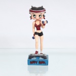 BRINQUEDO / COLECIONISMO. Estatueta decorativa colecionável em resina policromada figurando a personagem Betty Boop "Praticando Aeróbica". Coleção oficial licenciada, final do século XX ou início do século XXI. A icônica personagem surge representada em pé sobre uma plataforma de step cinza e preta, com o pé direito elevado em movimento de subida. Apresenta-se com seus característicos grandes olhos verdes e brincos de argola na cor vermelha, trazendo a cabeça cingida por uma faixa de suor(headband) nas cores vermelha e branca. Traja conjunto esportivo composto por top de alças em tom vermelho-grená metálico e shorts curto  na cor preta. Segura um par de halteres azuis, um em cada mão, mantendo o braço esquerdo flexionado à frente do tórax e o braço direito estendido junto ao corpo. Calça meias brancas e tênis esportivos vermelhos com solado branco; fixada sobre base retangular texturizada azul-turquesa. Na face frontal da base, inscrição em relevo com o nome da personagem "BETTY BOOP", destacado em esmalte vermelho com contorno preto. No reverso da base, apresenta inscrição de direitos autorais e licenciamento oficial gravada: "KFS/FS TM Hearst/FS". Medidas: Alt.: x Comp.: x Larg.: cm.