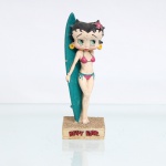 BRINQUEDO / COLEFCIONISMO. Estatueta decorativa colecionável em resina policromada figurando a personagem Betty Boop vestida como "Surfista". Coleção oficial licenciada, final do século XX ou início do século XXI. A icônica personagem surge representada em pé sobre base retangular texturizada em tom ocre-claro que simula uma superfície de areia de praia. Apresenta-se descalça, em posição frontal, ostentando seus tradicionais grandes olhos verdes e brincos de argola amarelos, trazendo o cabelo ornamentado por uma flor de hibisco na cor rosa com miolo amarelo na lateral esquerda. Traja biquíni na cor rosa com amarrações e debruns em azul-turquesa. Com o braço direito estendido para trás, segura e apoia uma grande prancha de surfe posicionada verticalmente ao seu lado;  face frontal da base, inscrição em relevo com o nome da personagem "BETTY BOOP", destacado em esmalte vermelho com contorno preto. No reverso da prancha de surfe, destaca-se aplicação de logotipo estilizado e colorido com a inscrição "Betty Boop". No reverso da base, apresenta inscrição de direitos autorais e licenciamento oficial gravada: "FS/FS TM Hearst/FS". Medidas: Alt.: x Comp.: x Larg.: cm.
